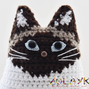 A crocheted cat toy with brown and white  fur. The cat has blue eyes and a brown nose. The toy is sitting on a white surface. The text 'ZOLAYKA knitting and crochet with love' is visible at the bottom of the image.