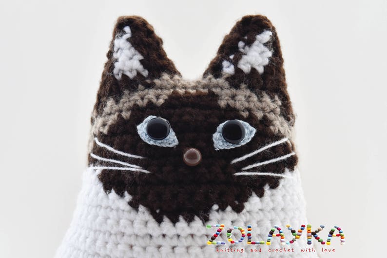 A crocheted cat toy with brown and white  fur. The cat has blue eyes and a brown nose. The toy is sitting on a white surface. The text 'ZOLAYKA knitting and crochet with love' is visible at the bottom of the image.