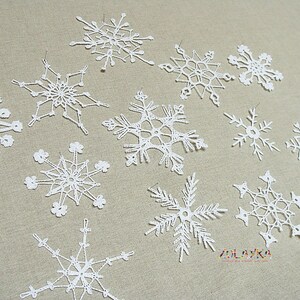 May include: A collection of twelve white crocheted snowflakes, each with a unique design, arranged on a beige fabric background.