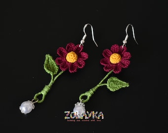 May include: Crocheted flower earrings with a burgundy flower, yellow center, green leaves, and a white pearl accent.  The earrings are on a black background.
