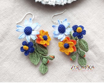 May include: A pair of handmade crochet earrings featuring a floral design. The earrings showcase blue, orange, and yellow flowers with green leaves. The earrings are on a light-colored fabric background. The text "ZOLAYKA" is visible.