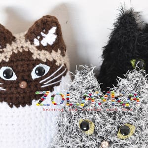 Three crocheted cat toys. One cat is brown and white, one is black, and one is gray with white fur. The gray cat toy has black eyes and a pink nose. The black cat toy has green eyes and a gray nose. The brown and white cat toy has brown eyes and a brown nose. The text 'ZOLANKA knitting and crochet with love' is visible in the image.