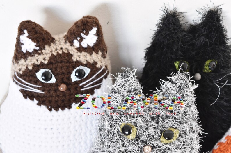 Three crocheted cat toys. One cat is brown and white, one is black, and one is gray with white fur. The gray cat toy has black eyes and a pink nose. The black cat toy has green eyes and a gray nose. The brown and white cat toy has brown eyes and a brown nose. The text 'ZOLANKA knitting and crochet with love' is visible in the image.
