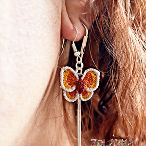 May include: Crocheted butterfly earrings with orange, yellow, white, and red accents hanging from silver hooks. The earrings are worn by a person with long brown hair.