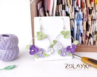 May include: Crocheted flower earrings with purple, white, and green flowers on silver hoops. The earrings are displayed on a white card with the text "ZOLAYKA knitting and crochet with love".