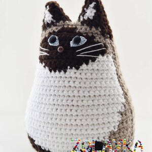 A crocheted cat toy with brown, white, and beige yarn. The cat has blue eyes and a brown nose. The cat is sitting on a white surface. The text 'ZOLAYKA knitting and crochet with love' is on the bottom of the image.