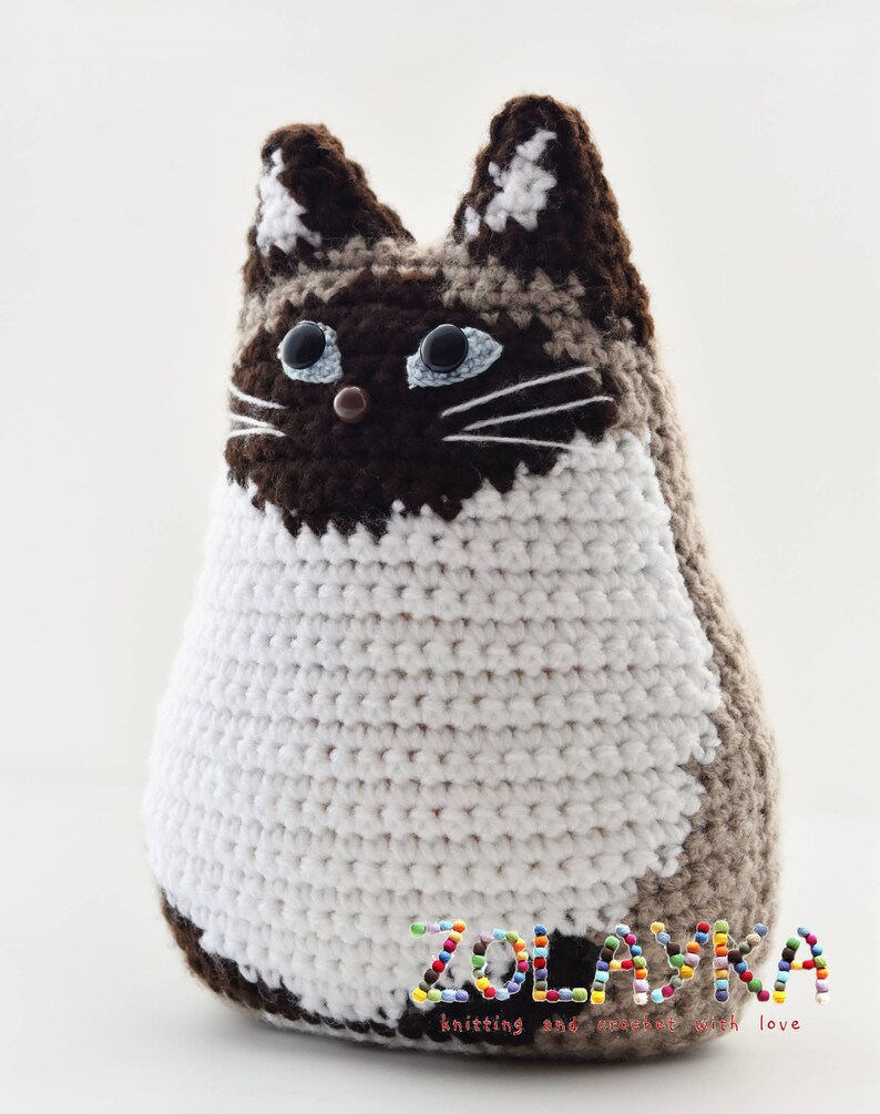 A crocheted cat toy with brown, white, and beige yarn. The cat has blue eyes and a brown nose. The cat is sitting on a white surface. The text 'ZOLAYKA knitting and crochet with love' is on the bottom of the image.