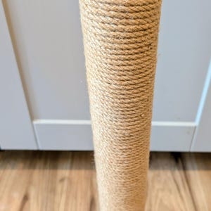 May include: A cylindrical cat scratching post. The post is wrapped in tightly wound, natural-colored rope. The top has a black circular base with pre-drilled holes. The post stands vertically on a wooden floor.
