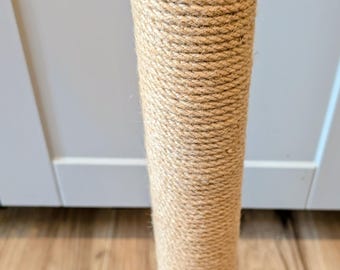 May include: A cylindrical cat scratching post. The post is wrapped in tightly wound, natural-colored rope. The top has a black circular base with pre-drilled holes. The post stands vertically on a wooden floor.