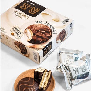 Pode incluir: Uma caixa de bolos de creme Lotte Mongshell Original com cobertura de chocolate e recheio de creme. A caixa apresenta imagens dos bolos e texto coreano. Vários bolos embalados individualmente e um prato de madeira com bolos também são visíveis.