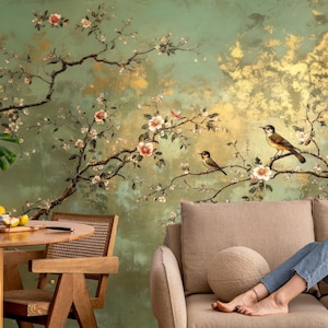 May include: A decorative wall mural featuring a nature-inspired design. The mural showcases a green and gold background with branches of flowering trees and perched birds. A round wooden table and a beige sofa are in the foreground.