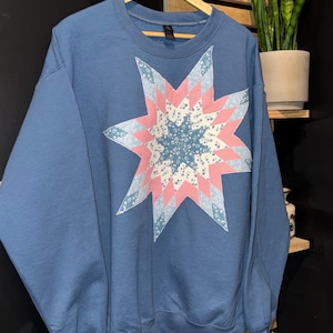 May include: A blue sweatshirt with a large star design on the front. The star is made of various fabric patterns in pink, blue, and white. The sweatshirt is hanging on a wooden hanger.