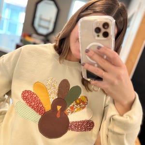 May include: A light beige sweatshirt featuring a colorful turkey applique. The turkey has a brown body, a red wattle, and a fan of patterned feathers in shades of red, yellow, green, and floral prints. The sweatshirt has long sleeves.