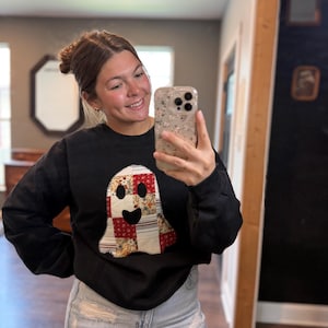 Op de afbeelding: Zwart sweatshirt met een patchwork spookontwerp met rode, witte en bloemenstoffen. De geest heeft een lachend gezicht. Het sweatshirt wordt gecombineerd met een lichtblauwe jeans.
