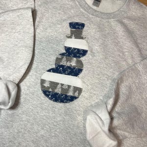 May include: A light gray sweatshirt featuring a snowman applique. The snowman is constructed from various patterned fabrics, including blue with white snowflakes, gray with trees, and white stripes. The applique is centered on the chest.