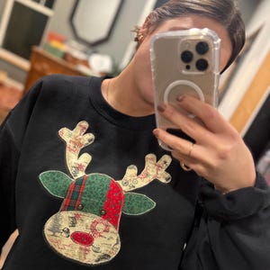 Op de afbeelding: Zwart sweatshirt met een rendierontwerp. Het rendier heeft groene en rode geruite geweien en een patroon gezicht met een rode neus. De sweatshirt heeft een ronde hals en lange mouwen.