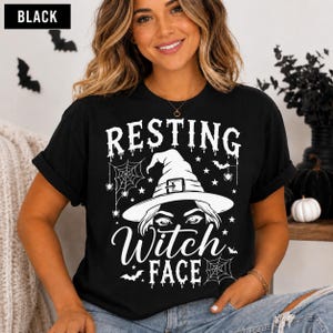 May include: A black t-shirt featuring a white graphic print with the text "RESTING WITCH FACE". The design includes a witch hat, spider webs, stars, and bats. The text "RESTING" and "FACE" appear in a dripping font style above and below the hat.