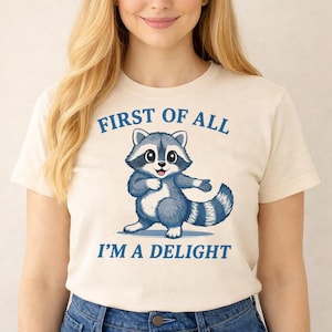 Könnte beinhalten: Cremefarbenes T-Shirt mit einem Cartoon-Waschbären in Blau und Grau. Der Waschbär lächelt und trägt den blauen Text "FIRST OF ALL I'M A DELIGHT" über und unter der Illustration.