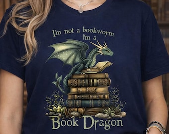 May include: Navy blue t-shirt featuring a green dragon perched atop a stack of books. The shirt has the text "I'm not a bookworm I'm a Book Dragon" in a decorative font. The dragon has detailed wings and scales.