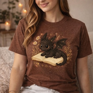 以下が含まれることがあります： 漫画のドラゴンが描かれた茶色のTシャツ。大きな目と星型の翼を持つ黒いドラゴンが、開いた本の上にいます。本は黄ばんだページで、デザインには白と黄色の花が含まれています。
