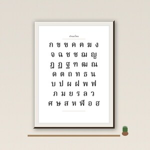 Op de afbeelding: Een ingelijste print met Thaise letters in zwart op een witte achtergrond. De print hangt aan een lichtgekleurde muur, met rechts een kleine plant in een bruine pot. De lijst heeft een neutrale kleur.