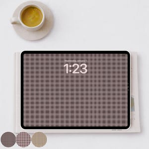 Puede incluir: Una tableta digital con un patrón de cuadros marrones en la pantalla, que muestra la hora 1:23. Una pequeña taza de café está sobre un platillo en la esquina superior izquierda. La tableta descansa sobre un libro con tres muestras de color en la parte inferior.