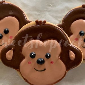 May include: Three decorated sugar cookies shaped like monkey faces. The cookies are brown, pink, and black with white icing. The cookies are on a white surface.