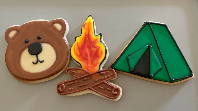 K&ouml;nnte beinhalten: Drei verzierte Zuckerkekse: ein brauner B&auml;r, ein gr&uuml;nes Zelt und ein Lagerfeuer mit orangefarbenen Flammen und braunen Holzscheiten.