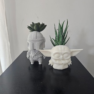 May include: Two character-shaped planters. One is a gray, speckled, helmeted figure holding a dark green succulent. The other is a white, wide-eared, small creature figure holding a spiky green succulent. Both are molded, matte-finish containers.