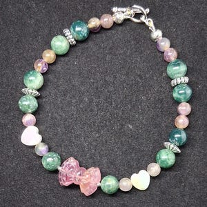 May include: A beaded bracelet featuring a variety of colorful beads. The bracelet includes green, pink, and purple beads, along with heart-shaped and bow-shaped accents. The bracelet is secured with a silver clasp.