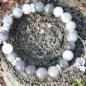 May include: A beaded bracelet featuring gray and white round stones. Silver-toned charms include stars, moons, and decorative accents. The bracelet is displayed on a textured, brown surface.