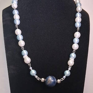 May include: A necklace featuring a combination of white, light blue, and dark blue beads. The necklace has a central dark blue stone and is displayed on a black mannequin. The beads are strung on a silver chain.