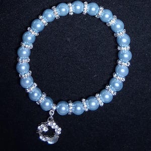May include: A light blue beaded bracelet with silver-colored rhinestone accents. The bracelet features a charm with a crescent moon design, also adorned with rhinestones. The beads are round and evenly spaced.