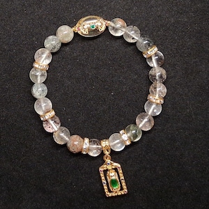 May include: A beaded bracelet featuring translucent, speckled clear quartz beads interspersed with gold-toned spacers encrusted with small clear crystals. The bracelet includes a central oval bead and a rectangular gold-toned pendant with a green stone.