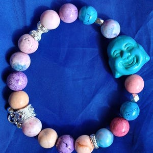 May include: A beaded bracelet featuring a turquoise Buddha head and a variety of colorful beads. The beads are in shades of pink, purple, blue, and peach, with silver-toned spacers. The bracelet is on a blue background.
