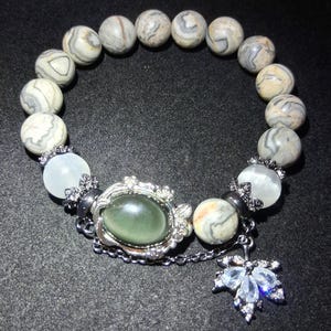 May include: A beaded bracelet featuring round, patterned gray and cream-colored beads. The bracelet includes a central oval green stone set in a silver-toned frame, and a dangling leaf charm with clear stones.