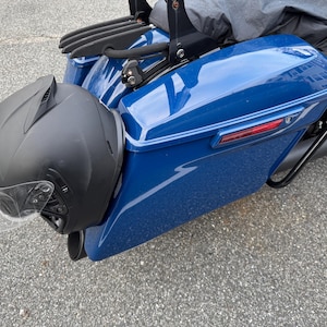 May include: A black motorcycle helmet with a clear visor is mounted on the side of a blue motorcycle. The motorcycle has a black luggage rack and a black exhaust pipe. The image is taken outdoors.