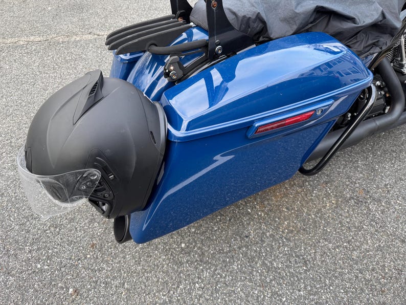 May include: A black motorcycle helmet with a clear visor is mounted on the side of a blue motorcycle. The motorcycle has a black luggage rack and a black exhaust pipe. The image is taken outdoors.