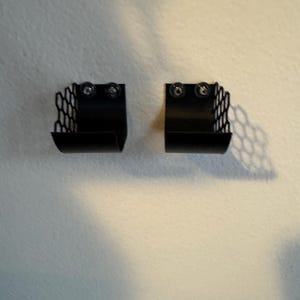 Puede incluir: Dos cestas de almacenamiento de metal negro montadas en la pared. Cada cesta tiene un patr&oacute;n de panal en los lados y se asegura con dos tornillos. Las cestas est&aacute;n dise&ntilde;adas para guardar objetos peque&ntilde;os y est&aacute;n montadas en una pared blanca.
