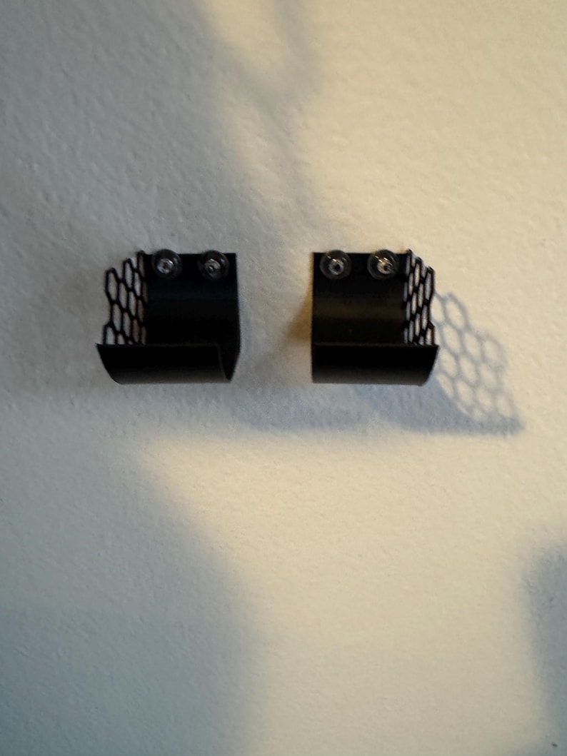 Puede incluir: Dos cestas de almacenamiento de metal negro montadas en la pared. Cada cesta tiene un patr&oacute;n de panal en los lados y se asegura con dos tornillos. Las cestas est&aacute;n dise&ntilde;adas para guardar objetos peque&ntilde;os y est&aacute;n montadas en una pared blanca.