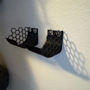 Puede incluir: Estante negro, montado en la pared, impreso en 3D, con un dise&ntilde;o de panal. El estante tiene una forma curva y est&aacute; dise&ntilde;ado para guardar objetos peque&ntilde;os. Dos objetos redondos y transparentes est&aacute;n unidos al estante.