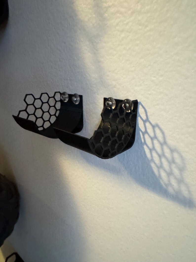 Puede incluir: Estante negro, montado en la pared, impreso en 3D, con un dise&ntilde;o de panal. El estante tiene una forma curva y est&aacute; dise&ntilde;ado para guardar objetos peque&ntilde;os. Dos objetos redondos y transparentes est&aacute;n unidos al estante.