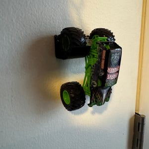 Puede incluir: Un cami&oacute;n monstruo de juguete verde y negro con neum&aacute;ticos negros grandes. El cami&oacute;n tiene las palabras "Grave Digger" impresas en el lateral. El cami&oacute;n est&aacute; boca abajo y montado en una pared blanca.