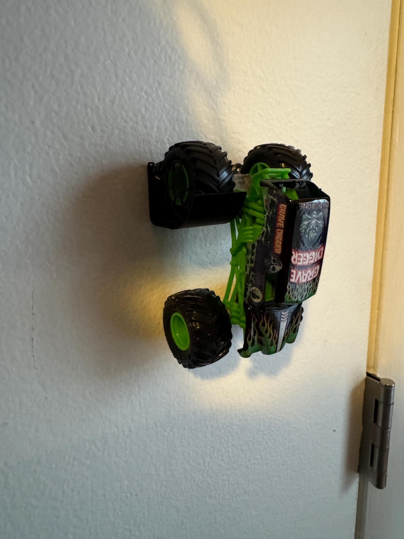 Puede incluir: Un cami&oacute;n monstruo de juguete verde y negro con neum&aacute;ticos negros grandes. El cami&oacute;n tiene las palabras "Grave Digger" impresas en el lateral. El cami&oacute;n est&aacute; boca abajo y montado en una pared blanca.