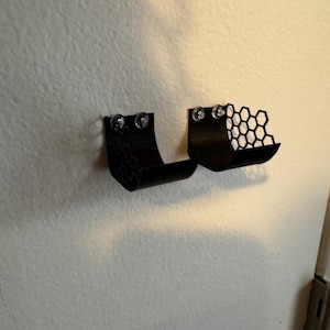 Puede incluir: Dos estantes negros montados en la pared con un dise&ntilde;o de panal. Cada estante se fija a la pared con dos tornillos transparentes. Los estantes tienen una forma curva, dise&ntilde;ados para sostener objetos peque&ntilde;os. El fondo es una pared lisa blanquecina.