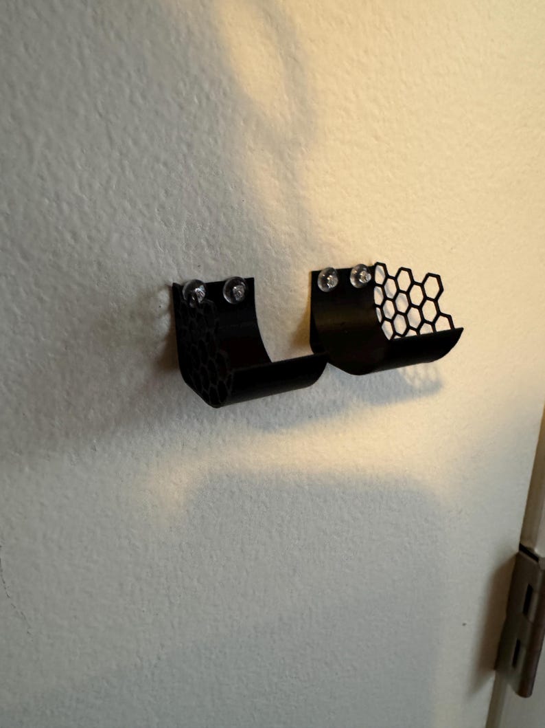 Puede incluir: Dos estantes negros montados en la pared con un dise&ntilde;o de panal. Cada estante se fija a la pared con dos tornillos transparentes. Los estantes tienen una forma curva, dise&ntilde;ados para sostener objetos peque&ntilde;os. El fondo es una pared lisa blanquecina.
