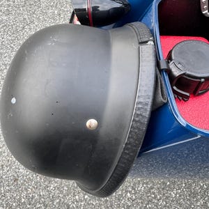 May include: A black motorcycle helmet with a textured strap is attached to a blue motorcycle. The helmet has a matte finish and a single silver rivet. The motorcycle has a red interior.