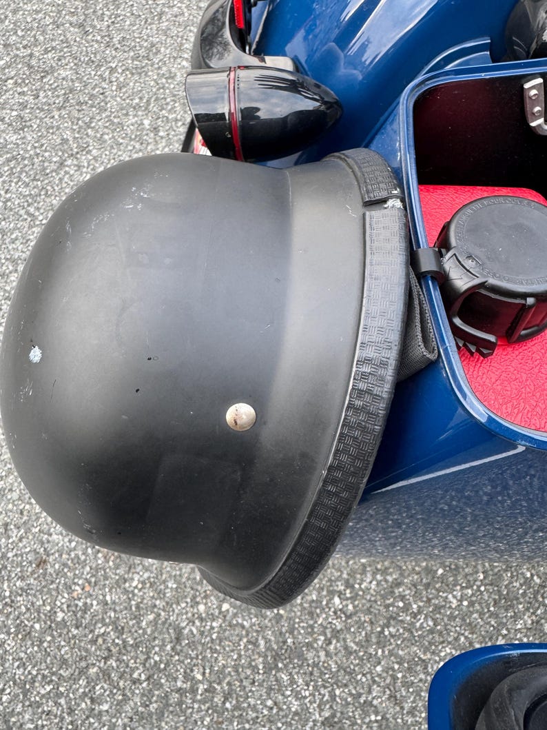 May include: A black motorcycle helmet with a textured strap is attached to a blue motorcycle. The helmet has a matte finish and a single silver rivet. The motorcycle has a red interior.