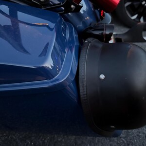 May include: A close-up of a black motorcycle helmet next to a dark blue vehicle. The helmet is matte black with a silver rivet. The vehicle has a glossy finish. A black wheel is visible in the background.