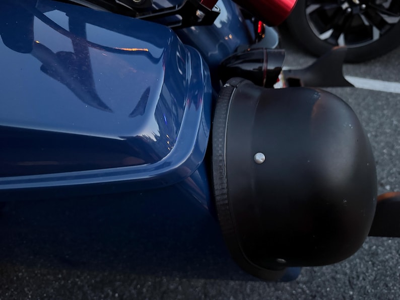 May include: A close-up of a black motorcycle helmet next to a dark blue vehicle. The helmet is matte black with a silver rivet. The vehicle has a glossy finish. A black wheel is visible in the background.